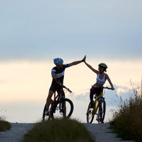 Sporty woman and man riding bicycles, having fun outside. Silhouettes of sportsmen highing five and posing on road in sunset time. Non urban scene.